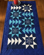 Dark blue tablerunner with white and blue pieced stars and embroidery.