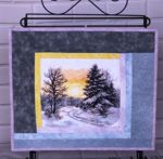 A small wall quilt with a stitch-out of a winter landscape in the center.