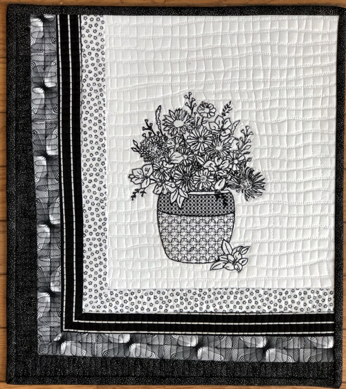 Finished quilt with black embroidery on the white background. The left and lower borders are pieced using 4 black-and-white fabrics. Black binding.