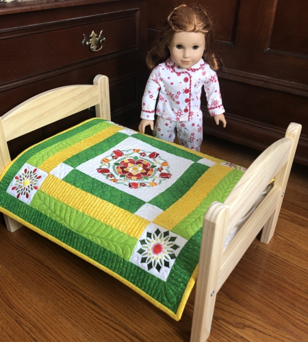 FInished small quilt in yellow and green colors with flower embroidery on a doll bed.