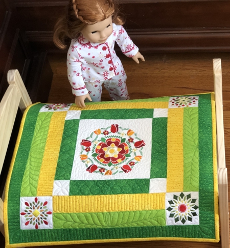 Finished small quilt in yellow and green colors with flower embroidery shown on a doll bed.