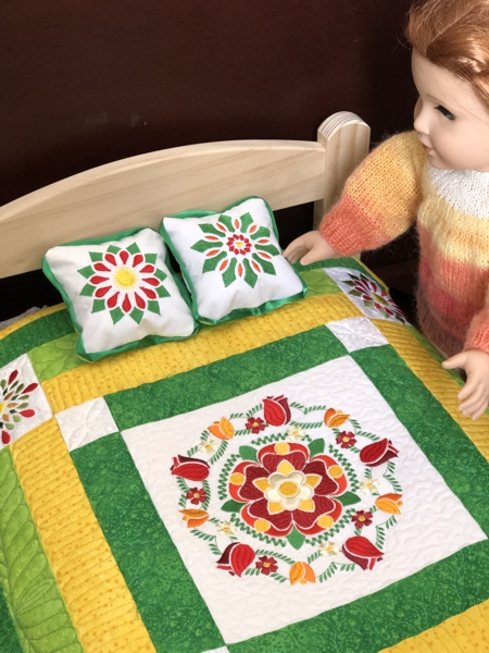 2 small pillows and finished small quilt in yellow and green colors with flower embroidery shown on a doll bed.