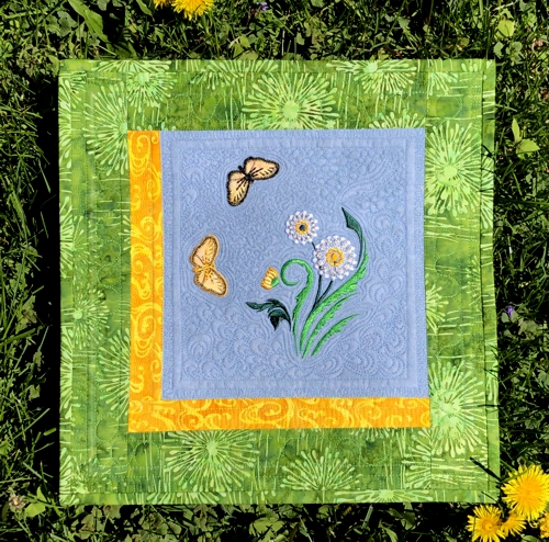 Small quilt with blue central part and embroidery of a dandelion and butterflies on it, yellow and green borders.