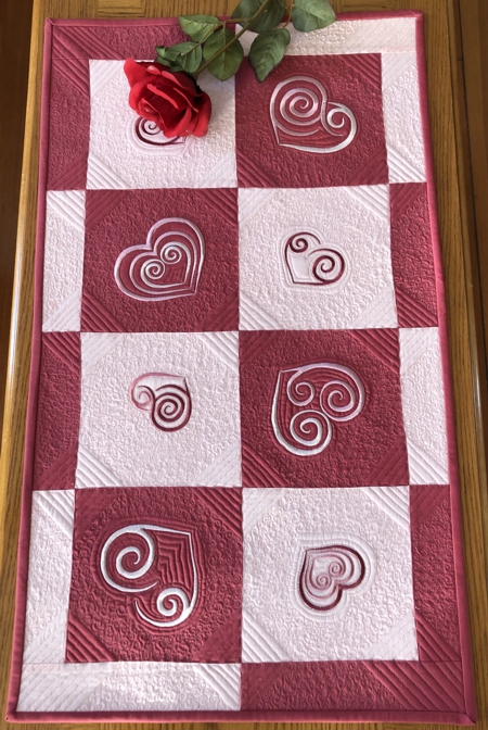 Finished quilted tablerunner with dark and pale pink squares and heart embroidery.