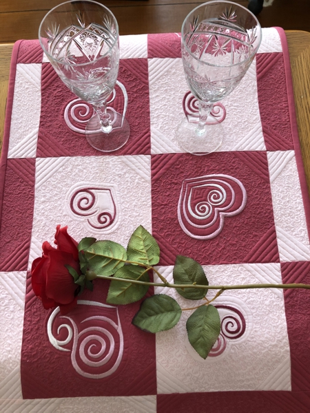 Finished quilted tablerunner with dark and pale pink squares and heart embroidery.