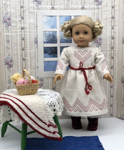 A doll in finished off-white dress with red embroidery on bodice, hem and sleeves.