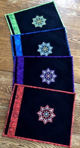 4 black place mats with embroidery and binding in jewel colors on a table.