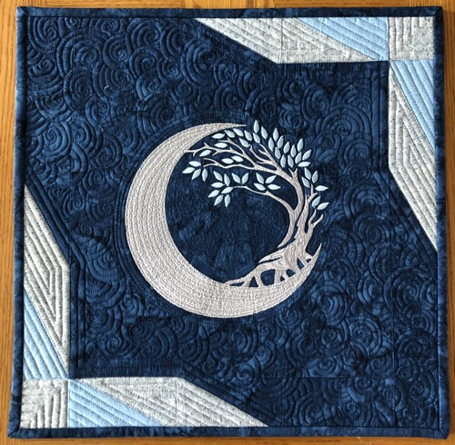 Small dark blue quilt with embroidery of a moon and tree in silver colors