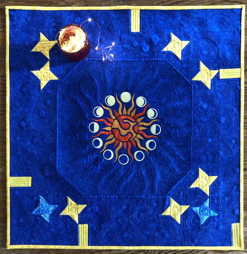 Royal blue quilted tabletopper with blocks of yellow stars and embroidery of the sun and moons in the center.