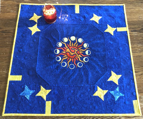 Royal blue quilted tabletopper with blocks of yellow stars and embroidery of the sun and moons in the center.