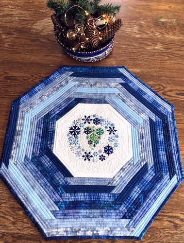 Quilted octagonal tabletopper made of blue scrap fabrics and snowflake embroidery in the center.