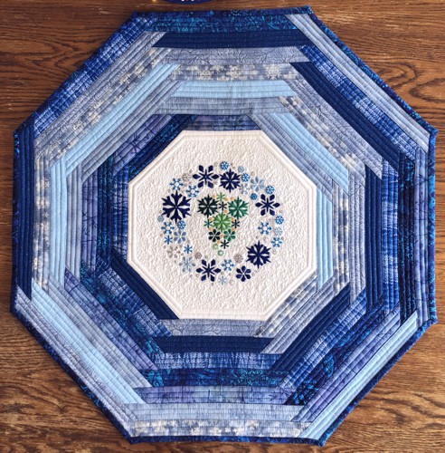 Quilted octagonal tabletopper made of blue scrap fabrics and snowflake embroidery in the center.