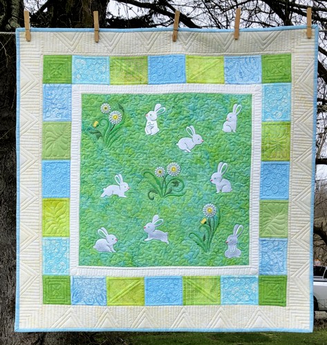 A small wall quilt for a nursery with embroidery of bunnies and dandelions in the central part, with 3 borders , all in green and blue shades.