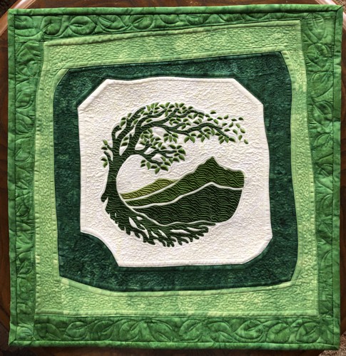 A small quilt with an embroidered green tree and mountains in the background, with 3 curved borders , all in shades of green.