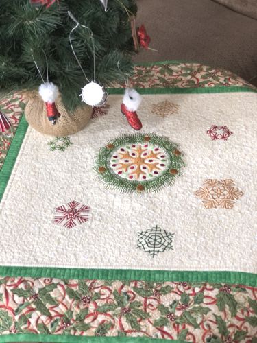 Quilted tabletopper with snowflake embroidery in the center and borders in Christmas patterned fabric.