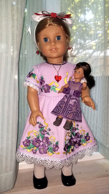 A 18-inch doll in a dress with flower embroidery holding a mini doll in lace dress.